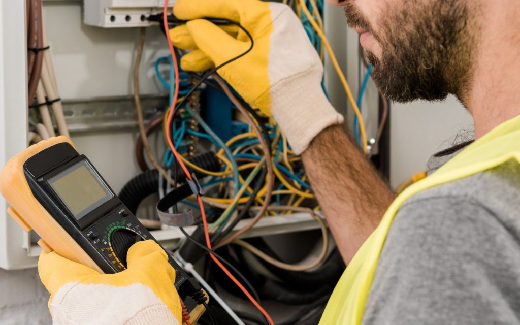 Electrician wearing yellow gloves and safety vest testing electrical wiring inside a switchboard with digital multimeter – Limitless NRG Solutions Brisbane & Sunshine Coast