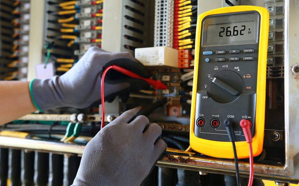 Electrician wearing gloves testing industrial switchboard voltage using a digital multimeter – Limitless NRG Solutions Brisbane & Sunshine Coast