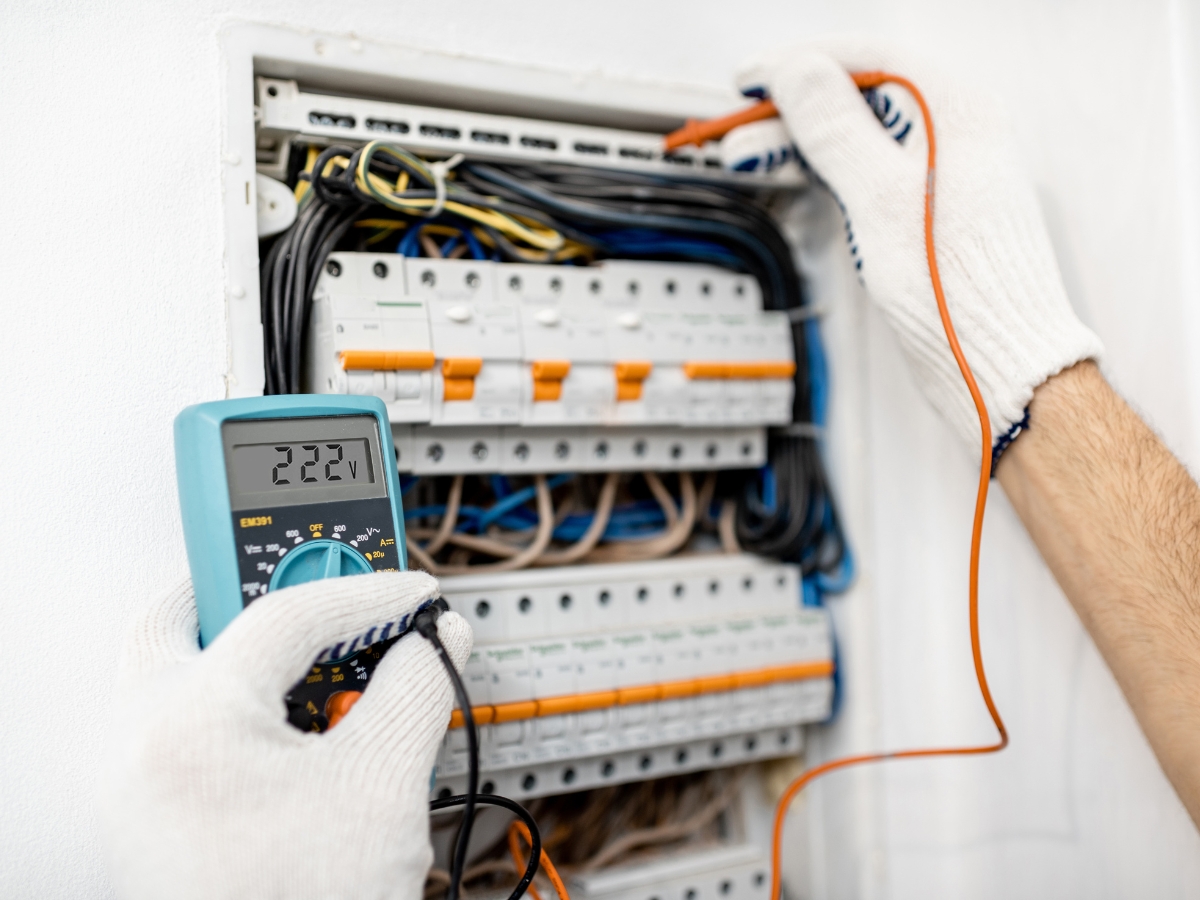 A person wearing white protective gloves uses a digital multimeter to measure voltage in a switchboard filled with wires and circuit breakers. The multimeter display shows a reading of 222.2 volts.
