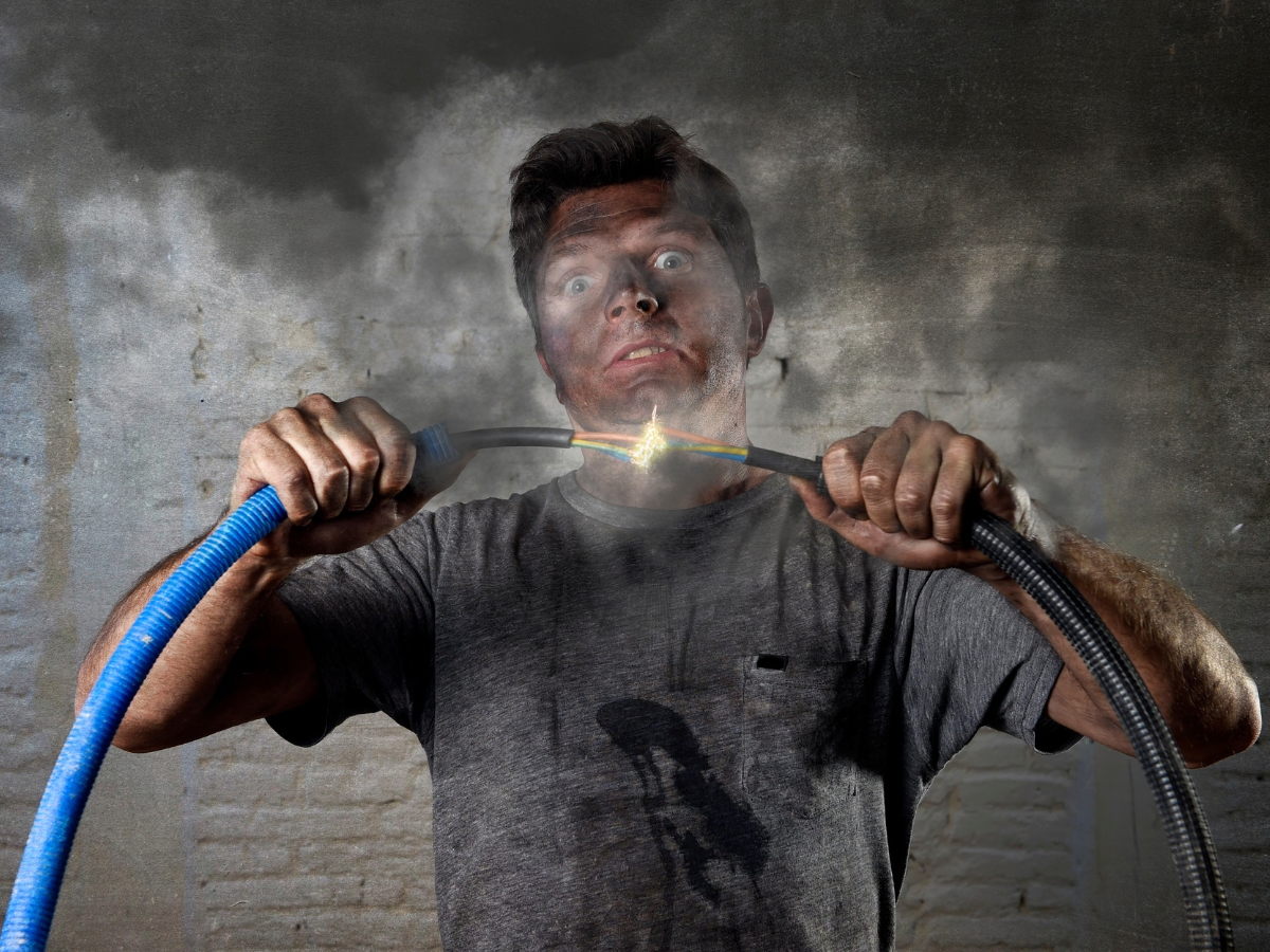 A man with a shocked expression holds two sparking electrical wires as smoke rises around him, highlighting the dangers of DIY electrical work. His face, clothes, and hands are smudged with dirt, suggesting an electrical mishap.