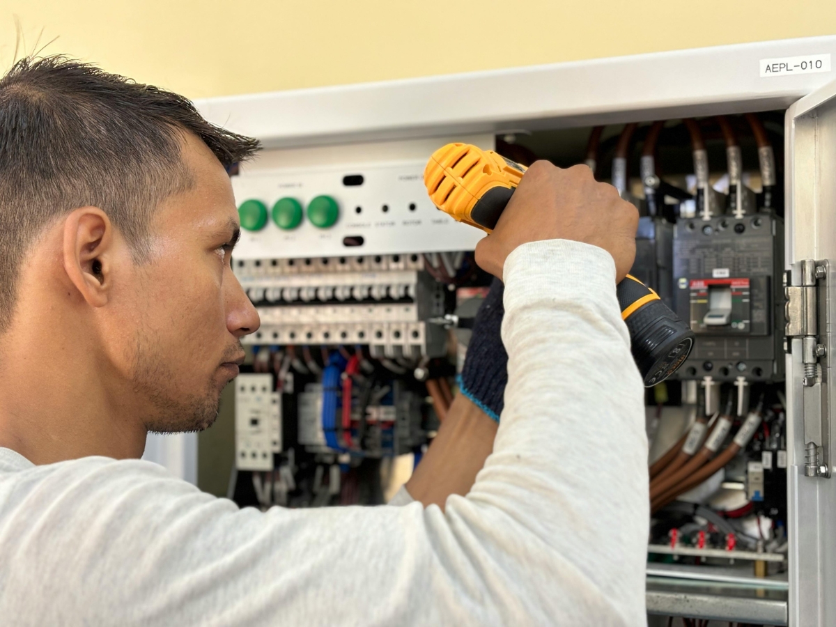 A man uses a yellow power drill to work on an open electrical switchboard filled with wires and switches. He is focused on making adjustments inside the panel.