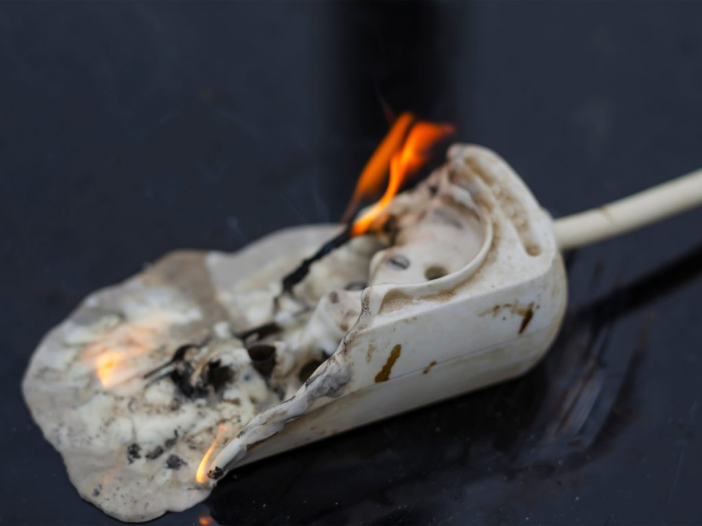 A partially melted and burnt power plug with visible flames sits on a dark surface, highlighting damage from intense heat and underscoring the importance of an electrical safety checklist.