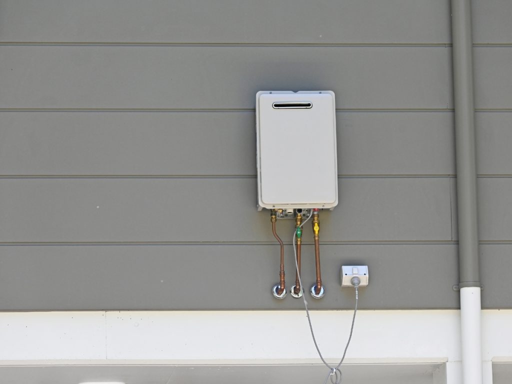 A tankless water heater is mounted on a grey wall with several pipes extending from its bottom; it appears to be part of a hot water system not working, as indicated by the nearby small control box connected by a wire.