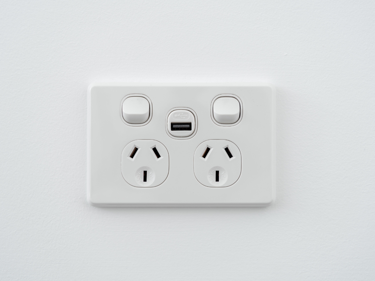 A white wall power point with two sockets, two on/off switches, and a USB charging port in the centre. This modern power point, featuring convenient USB sockets, is mounted on a plain white wall.