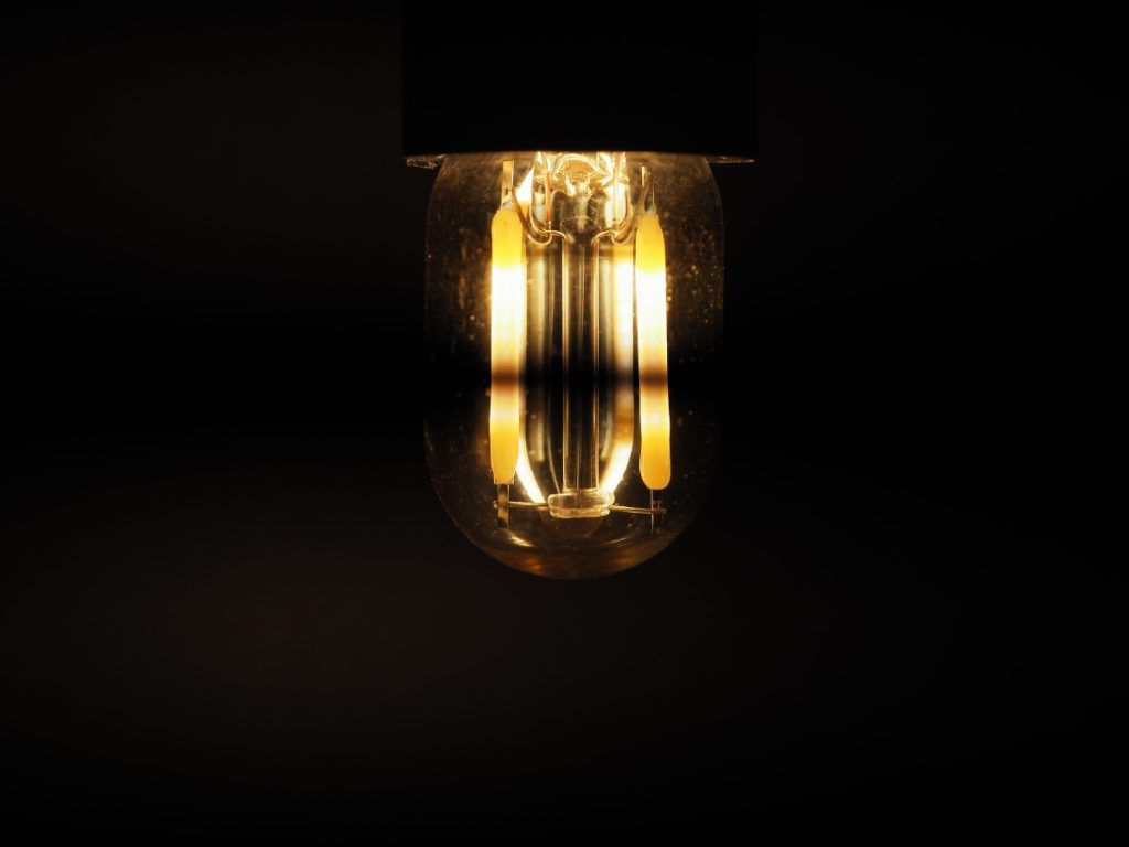 A glowing light bulb hangs against a dark, black background, its filament illuminated and casting a warm, soft glow, with faint lights flickering in the shadows.