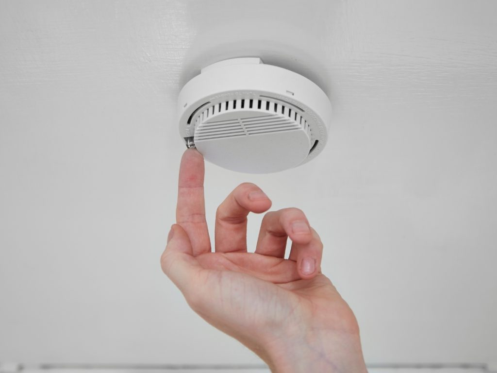 A hand reaching up to twist or test a white smoke alarm mounted on a white ceiling, ensuring safety and compliance ahead of the QLD Smoke Alarm Deadline.