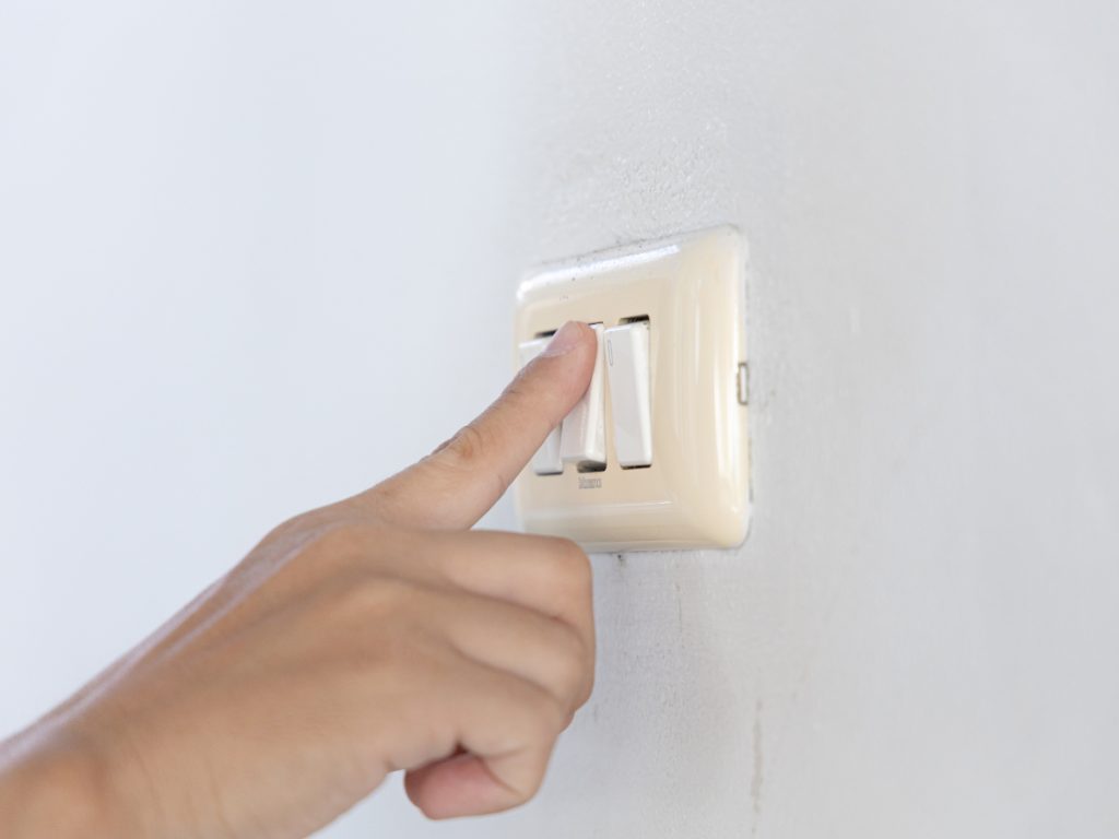 A person’s hand is pressing a white light switch on a white wall, possibly checking if a safety switch keeps tripping.