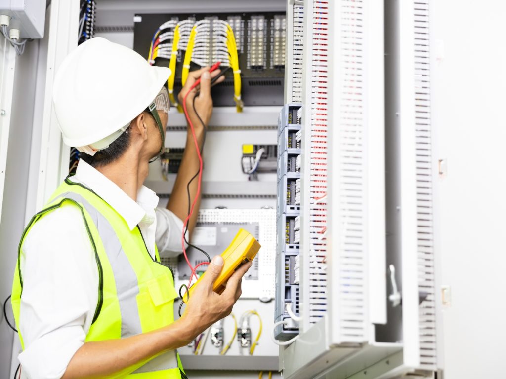 An electrician wearing a white helmet and yellow safety vest uses a multimeter to test wires inside an electrical switchboard. He is checking connections and equipment in an industrial setting.